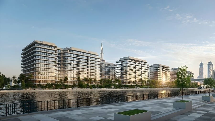 Dubai Creek Gardens seen across the water at sunset — low-rise residential wings along a landscaped waterfront promenade, with the Dubai skyline beyond
